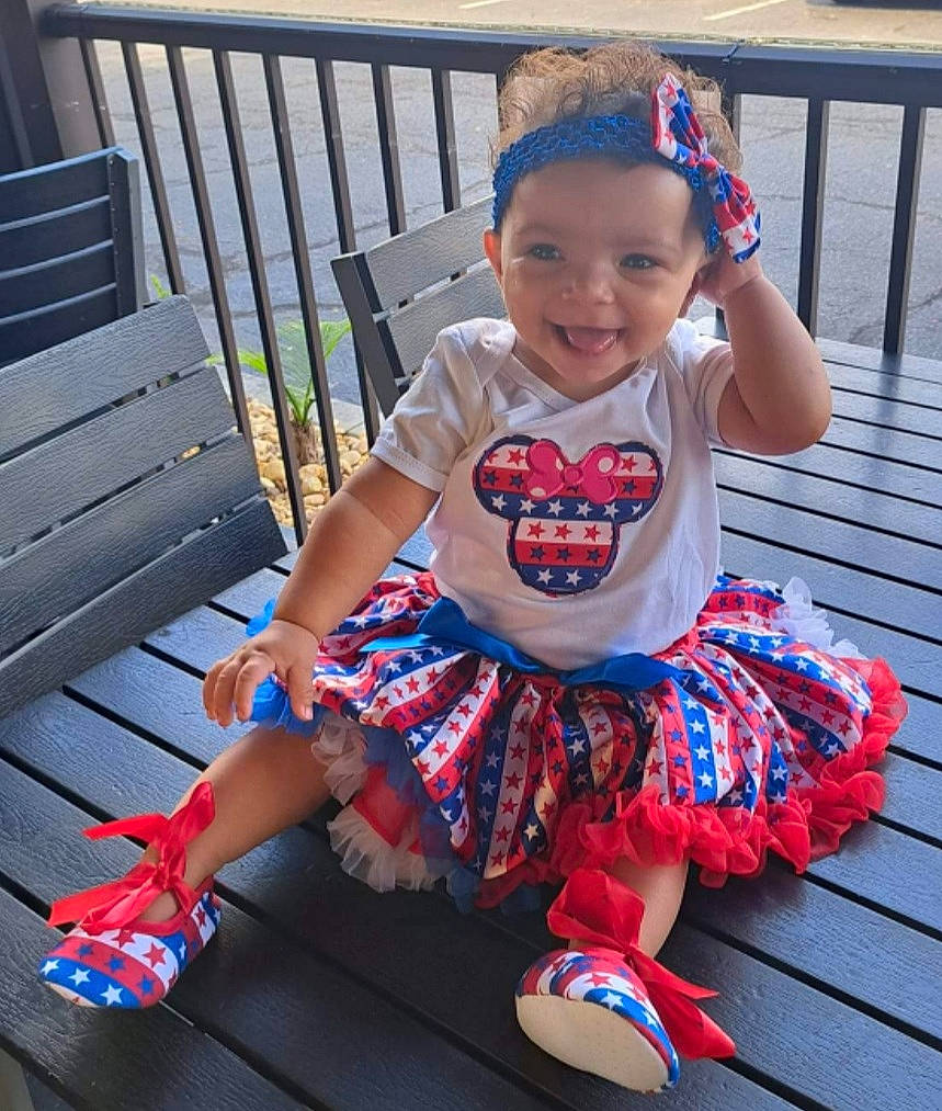 Gabriella is registered to the contest to win money with this photo: baby_toddler_clothing, child, electric_blue, fashion_accessory, foot, footwear, grass, happy, human_leg, joy, leisure, magenta, pattern, person, pink, sandal, sitting, sleeve, smile, t_shirt