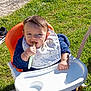 Julian a rejoint le concours — aidez-le/la à gagner de superbes lots ! baby, child, high_chair, bib, spoon, grass, outdoor, sunlight, blue_sweater, orange_chair, hand, face, smiling, young_child, person, feeding, happy, playful, daylight, casual