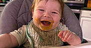 Emitte joined the competition — help win amazing prizes! baby, high_chair, smiling, messy_face, bib, food, kitchen, cabinet, microwave, countertop, person, child, happy, indoor, appliance, cooking, cute, infant, eating, expression