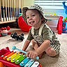 toddler, child, toy, xylophone, colorful, hat, striped_clothing, carpet, indoor, crib, window_blinds, smiling, playing, floor, toy_car, red_ball, furniture, happy, person, room