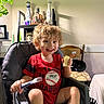 Malachai joined the competition — help win amazing prizes! child, toddler, high_chair, fork, banana, red_shirt, curly_hair, smiling, indoors, plant, decor, furniture, table, chair, happy, face, person, home, food, eating