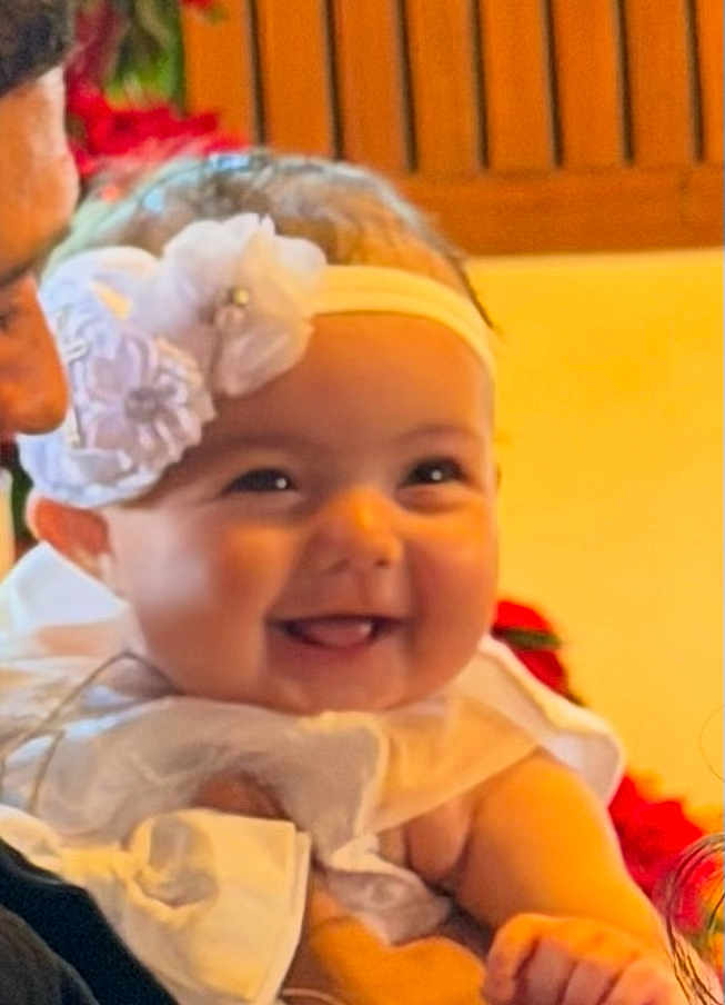 Eliana is registered to the contest to win money with this photo: baby, smiling, headband, flower, white_clothing, adult_hand, closeup, happy, infant, cute, portrait, indoor, warm_light, child, person, celebration, family, love, skin, face