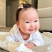Tia is registered to the contest to win money with this photo: baby, infant, smiling, face, hair_bow, blanket, carpet, living_room, white_clothing, cute, child, portrait, happy, lying_down, indoors, soft_texture, young_child, chubby_cheeks, bright_eyes, cozy