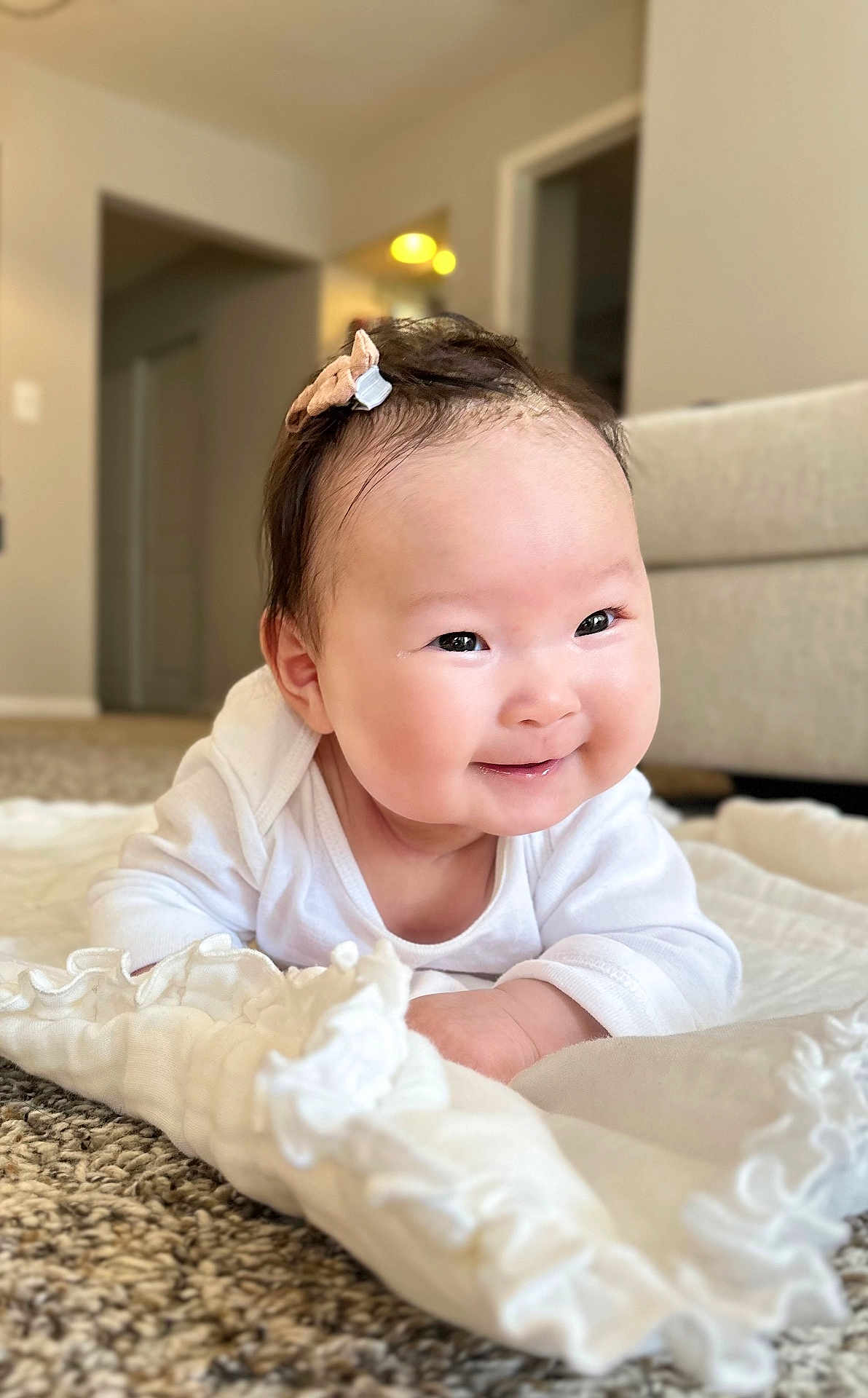 Tia is registered to the contest to win money with this photo: baby, infant, smiling, face, hair_bow, blanket, carpet, living_room, white_clothing, cute, child, portrait, happy, lying_down, indoors, soft_texture, young_child, chubby_cheeks, bright_eyes, cozy
