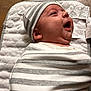 baby, infant, swaddle, hat, striped_clothing, quilted_mat, face, headwear, newborn, joyful, lying_down, closeup, skin, cute, child, expression, mouth_open, indoor, soft_textile, happy