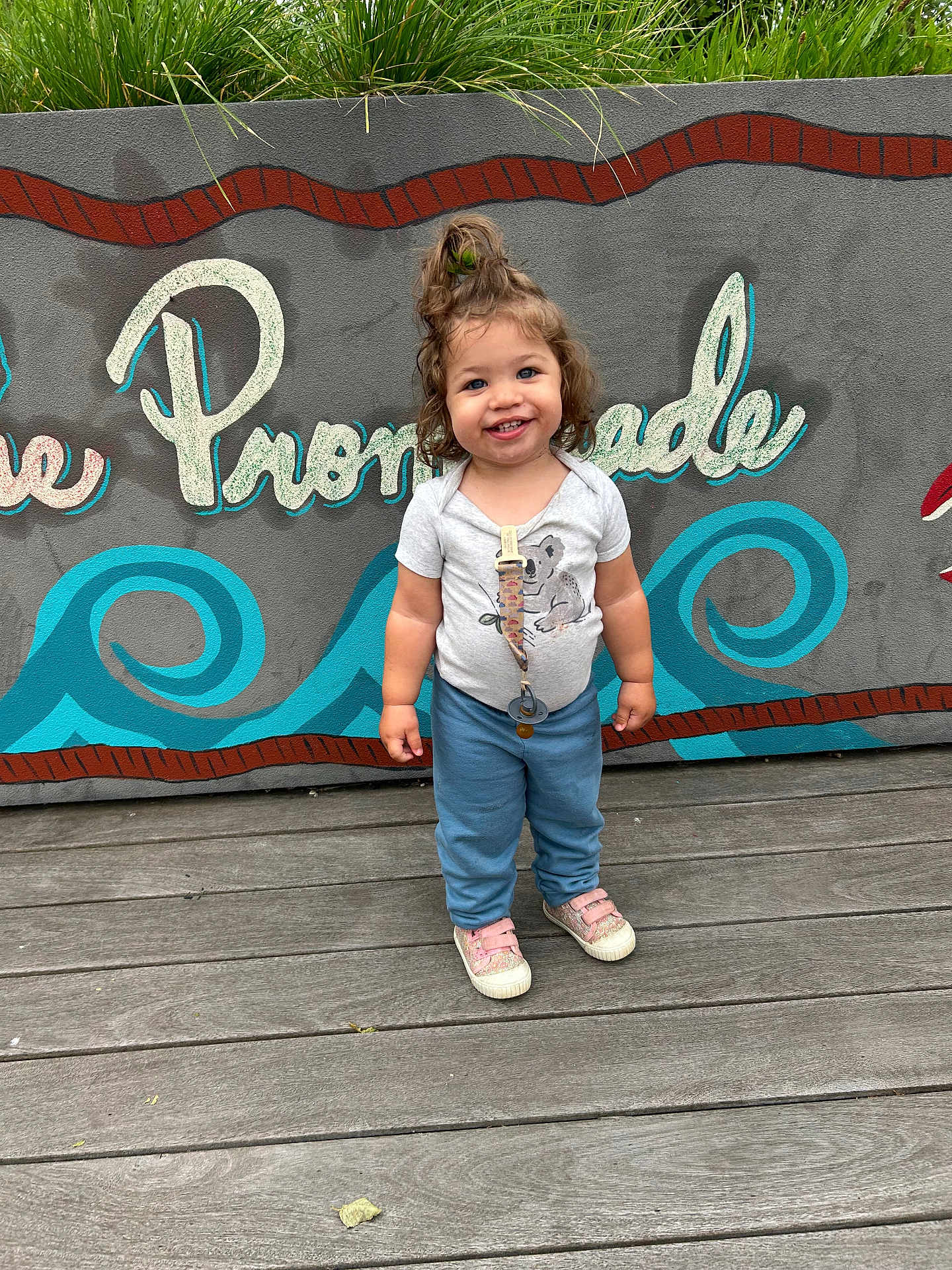 Jayley joined the competition — help win amazing prizes! toddler, child, smiling, standing, outdoor, wooden_floor, mural, wall_art, hair_tied, casual_clothing, sneakers, happy, person, daylight, blue_pants, grey_shirt, pacifier_clip, greenery, urban, playful