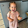 toddler, child, smiling, indoor, pink_dress, wet_hair, pigtails, carpet, cabinet, countertop, purple_shoes, patterned_clothing, holding_object, happy, person, floor, playful, cute, young_child, standing