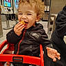 child, toddler, curly_hair, eating, snack, smiling, happy, restaurant, fast_food, shopping_cart, red_cart, seated, jacket, black_clothing, hand, food, portrait, indoor, food_court, family