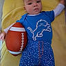 baby, child, infant, portrait, lying_down, football, sports, blue_shirt, lion_graphic, patterned_pants, yellow_blanket, playpen, indoor, face, looking_at_camera, serious_expression, hands, foot, soft_blanket, colorful_clothing