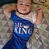 Brian joined the competition — help win amazing prizes! baby, child, infant, portrait, lying_down, smiling, indoor, cushion, pillow, blanket, blue_clothing, shirt, crown, text_on_clothing, geometric_pattern, bed, relaxed, face, arms, home