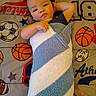 baby, infant, child, portrait, lying_down, blanket, swaddled, smiling, face, eyes, indoor, bed, bedding, sports_theme, basketball, football, stars, cozy, pointing, photography