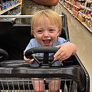 Zayne is registered to the contest to win money with this photo: toddler, child, shopping_cart, grocery_store, steering_wheel, happy, smiling, blond_hair, blue_eyes, person, tattoo, adult, aisle, products, shopping, retail, clothing, short_sleeves, floor, metal