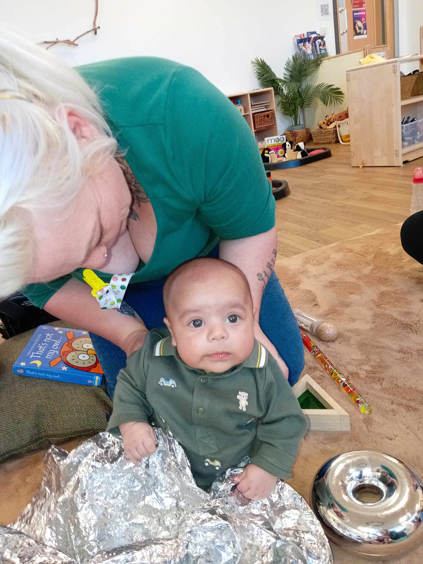 Tommy joined the competition — help win amazing prizes! baby, adult, green_shirt, blonde_hair, foil, toy, book, floor, carpet, indoor, person, child, wooden_toy, plant, sitting, curious, face, eyes, hand, room