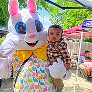 Ernest is registered to the contest to win money with this photo: baby, child, mascot, easter_bunny, costume, outdoor, tent, greenery, sunny, people, plaid_shirt, yellow_bow, colorful, holding, festival, event, smile, person, daylight, celebration