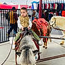 adult, animal, child, clothing_stall, crowd, fair_ride, fence, hat, jacket, outdoor_market, pavement, person, pet, pony, portrait, rope, saddle, sneakers, tent, toddler