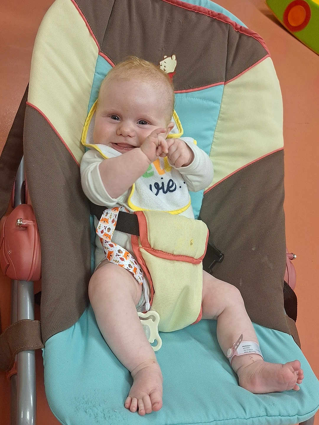 Astrid participe au concours pour gagner de l'argent avec cette photo : baby, smiling, clapping, baby_chair, bib, hospital_wristband, indoor, colorful, infant, cute, comfort, playful, child, person, foot, hand, face, happy, seat, fabric