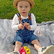 Aaron a rejoint le concours — aidez-le/la à gagner de superbes lots ! baby, child, hat, denim_overalls, toy, ball, blanket, grass, park, trees, sunlight, shoes, sitting, outdoor, nature, greenery, person, cute, infant, playful