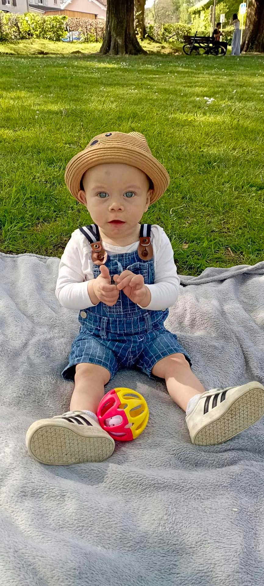 Aaron a rejoint le concours — aidez-le/la à gagner de superbes lots ! baby, child, hat, denim_overalls, toy, ball, blanket, grass, park, trees, sunlight, shoes, sitting, outdoor, nature, greenery, person, cute, infant, playful