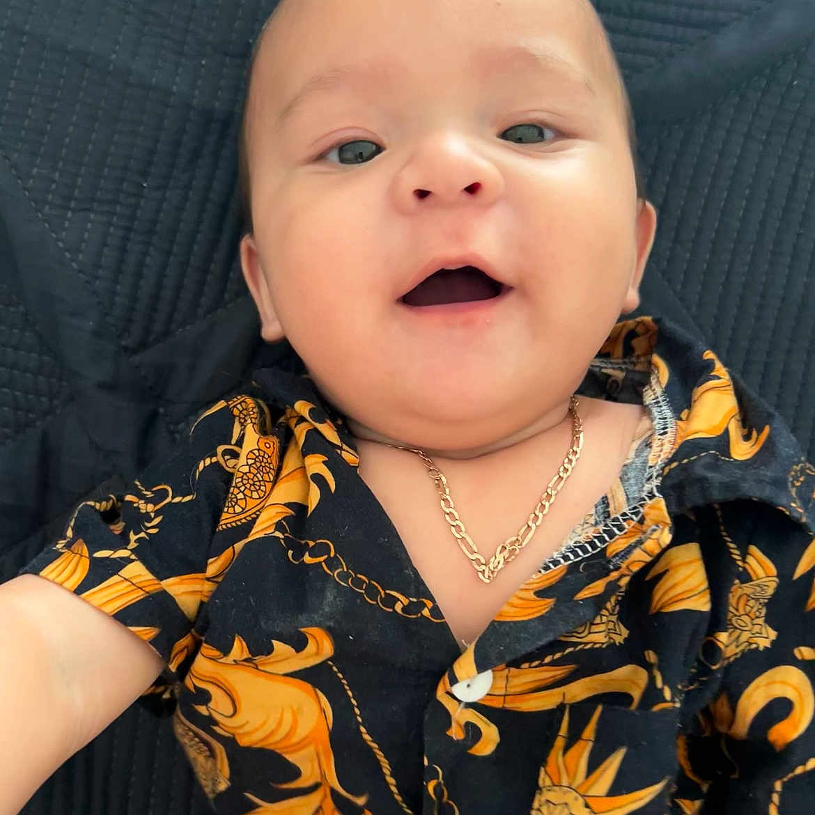 Julian Rodriguez is registered to the contest to win money with this photo: baby, black, child, closeup, cute, face, happy, indoors, infant, jewelry, lying_down, necklace, open_mouth, orange, patterned_clothing, person, portrait, shirt, smile, texture