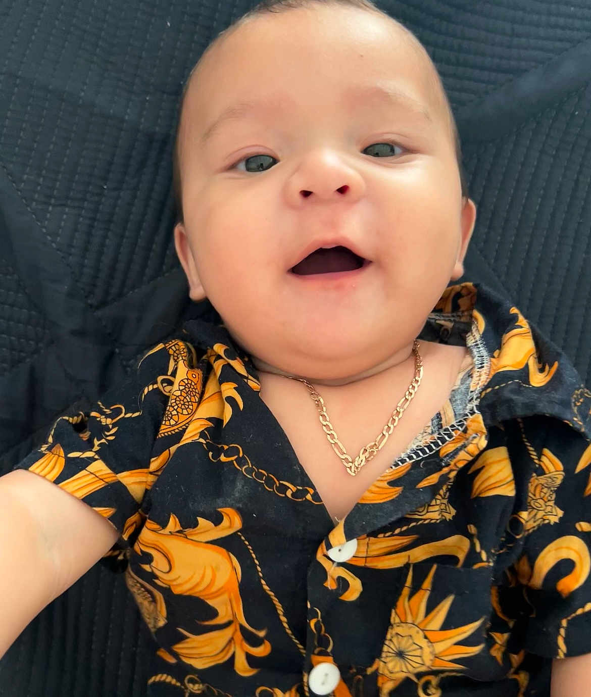 Julian Rodriguez is registered to the contest to win money with this photo: baby, child, smile, face, necklace, shirt, black, orange, patterned_clothing, open_mouth, lying_down, happy, cute, infant, person, portrait, jewelry, closeup, indoors, texture