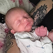 Caleb is registered to the contest to win money with this photo: newborn, baby, sleeping, tattoo, arm, onesie, space_theme, pillow, floral, plush_toy, dinosaur, hand, infant, person, cozy, indoor, closeup, resting, cute, portrait
