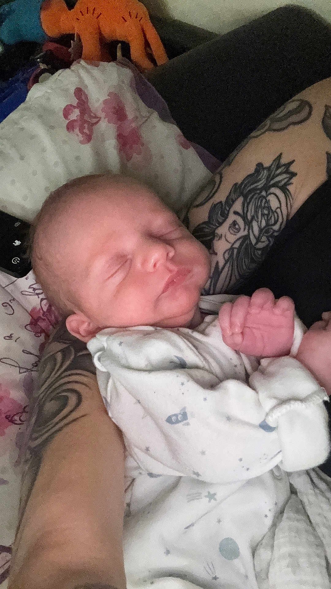 Caleb is registered to the contest to win money with this photo: newborn, baby, sleeping, tattoo, arm, onesie, space_theme, pillow, floral, plush_toy, dinosaur, hand, infant, person, cozy, indoor, closeup, resting, cute, portrait