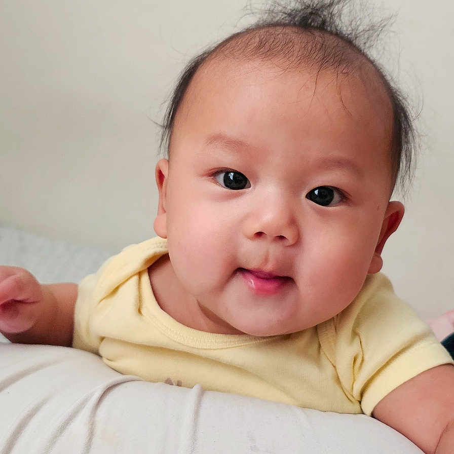 Nivaan Gurung is registered to the contest to win money with this photo: baby, infant, child, portrait, smiling, happy, face, eyes, tongue, hair, yellow_shirt, lying_down, pillow, bed, indoor, home, adult_hand, close_up, photography, cute