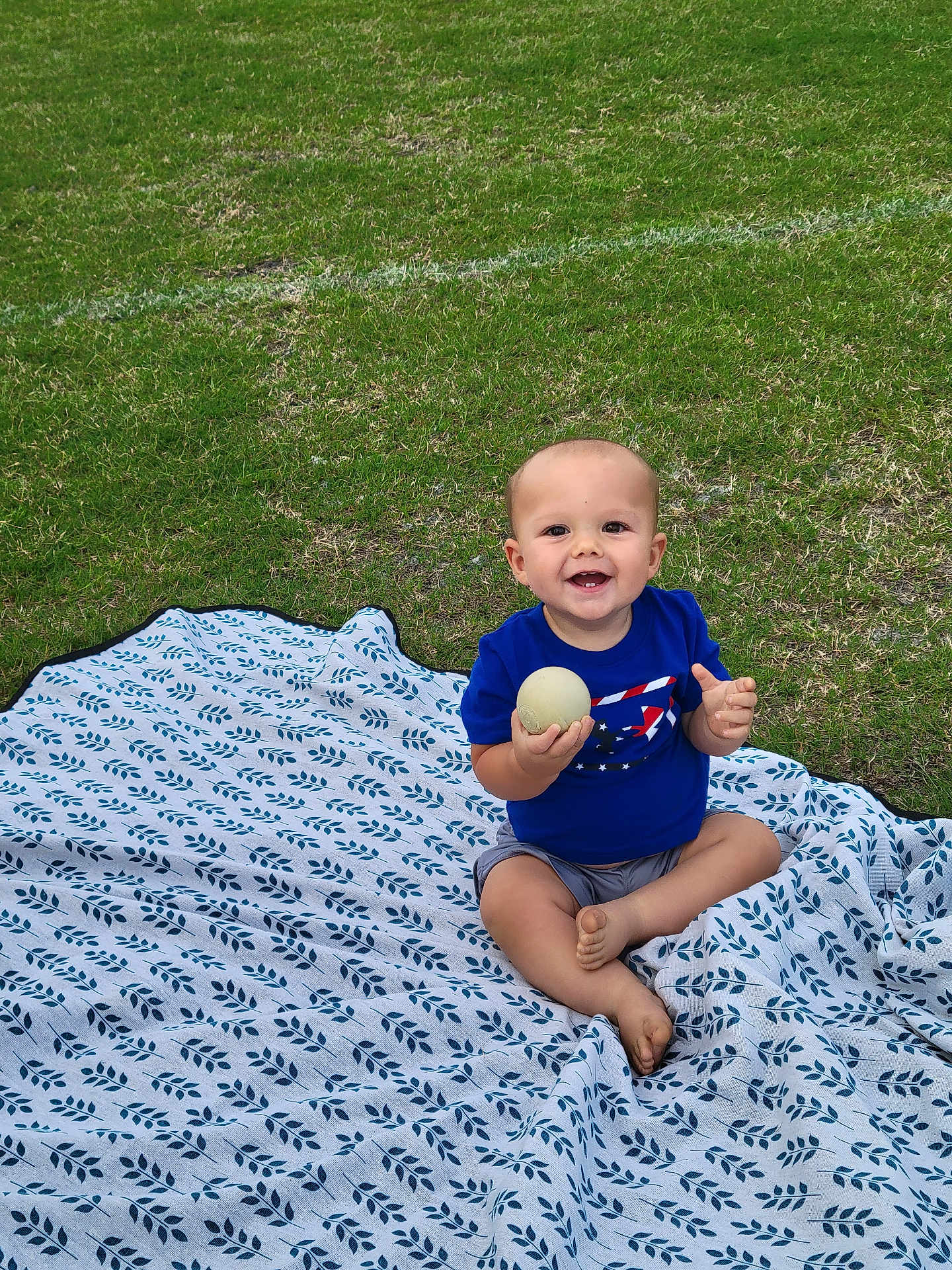 Camden is registered to the contest to win money with this photo: baby, ball, barefoot, blanket, blue_shirt, cheerful, child, cute, daylight, grass, greenery, happy, nature, outdoor, play, portrait, sitting, smiling, toddler, young_child