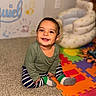baby, child, smiling, sitting, carpet, foam_puzzle_mats, striped_pants, green_top, indoor, inflatable_chair, handprints, wall, colorful, happy, cute, play_area, toddler, person, footwear, cozy