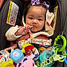 toddler, child, baby, car_seat, purple_bows, toys, colorful, finger_in_mouth, cute, clothing, baby_bottle, plush_toy, seat_belt, indoor, portrait, face, happy, small_hands, infant, baby_gear