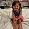 toddler, child, smiling, bed, white_bedspread, dress, stars, stripes, indoor, living_room, couch, pillow, curtain, window, foot, hair, person, happy, sitting, home