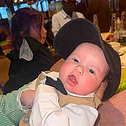 Anthony Melo is registered to the contest to win money with this photo: baby, child, infant, hat, cap, formal_wear, bow_tie, person, indoor, restaurant, table, glass, bottle, adult, blurred_background, face, skin, clothing, portrait, dining