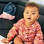 baby, child, sitting, couch, clothing, pink_top, socks, book, curly_hair, face, hand, indoor, furniture, expression, skin, person, cute, messy_face, bright_eyes, casual