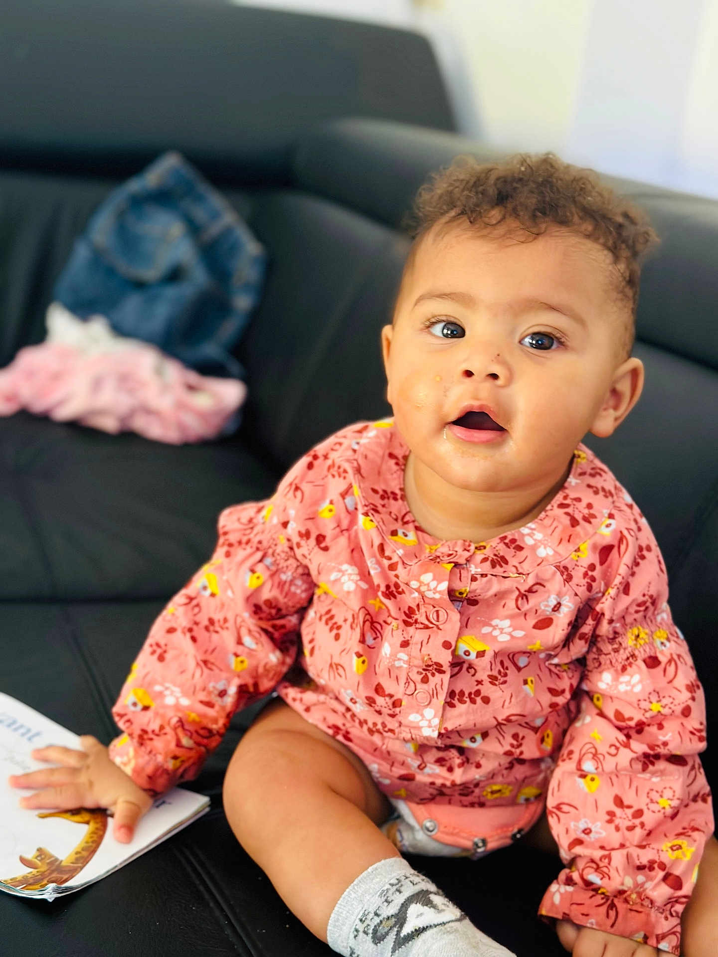 Jade participe au concours pour gagner de l'argent avec cette photo : baby, child, sitting, couch, clothing, pink_top, socks, book, curly_hair, face, hand, indoor, furniture, expression, skin, person, cute, messy_face, bright_eyes, casual