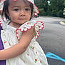 child, clothing, curious, cute, daylight, dress, face, fashion, hat, headwear, holding, nature, outdoor, parking_lot, person, smiling, summer, toddler, trees, young_child