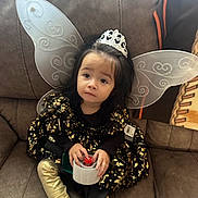 Scarlette joined the competition — help win amazing prizes! child, fairy_wings, tiara, dress, golden_leggings, tape_dispenser, couch, indoor, curly_hair, black_hair, decorative, seated, costume, young_child, playful, portrait, cute, fingertips, holding, expression