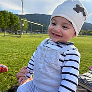 Aïden a rejoint le concours — aidez-le/la à gagner de superbes lots ! baby, child, smiling, outdoor, grass, field, hat, striped_clothing, person, happy, hands, nature, daylight, hill, casual_clothing, cute, infant, sitting, toothy_smile, playful