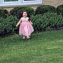 brick_wall, bushes, casual, child, curly_hair, daylight, flowering_plants, front_yard, girl, grass, happy, lawn, nature, outdoor, person, pink_dress, playful, running, smiling, window