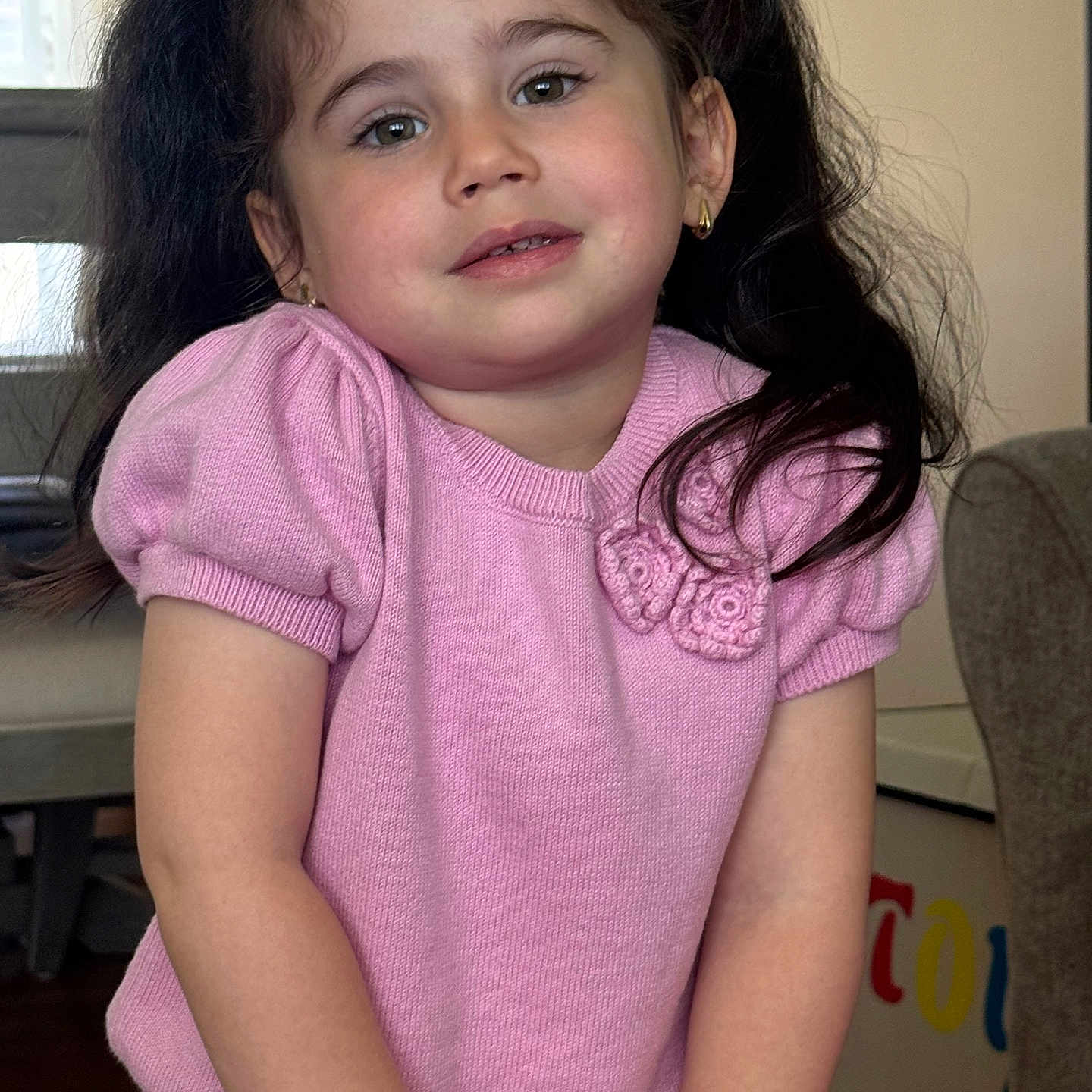 Snezhana is registered to the contest to win money with this photo: casual_clothing, child, colorful, cute, earrings, face, furniture, girl, hair, happy, home, indoor, person, pigtails, pink_sweater, portrait, skin, smile, toy_box, young_child