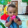 baby, child, toy, indoor, playmat, curly_hair, bib, colorful, nursery, plastic_toy, red_yellow_play_structure, floor, crib, door, wall_art, bird_mural, striped_shirt, basket, floor_mat, curious