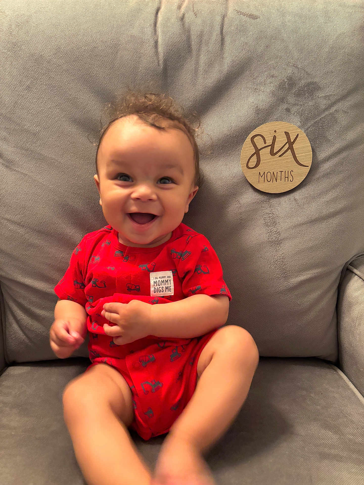 Ronan is registered to the contest to win money with this photo: baby, infant, smiling, red_onesie, curly_hair, gray_couch, wooden_sign, six_months, happy, cute, child, indoor, portrait, sitting, person, young_child, candid, joyful, expression, comfortable