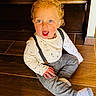 toddler, child, curly_hair, blue_eyes, suspenders, long_sleeve_shirt, patterned_pants, socks, wooden_floor, indoor, happy, smiling, sitting, cute, young_child, casual_clothing, portrait, person, flooring, bright