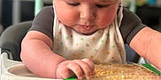 Heath is registered to the contest to win money with this photo: baby, infant, high_chair, toy, hand, foot, face, skin, clothing, bib, tape, table, indoors, child, cute, expression, sitting, furniture, focus, background_blur