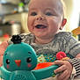 Heath is registered to the contest to win money with this photo: baby, smiling, toy, bird_toy, happy, child, indoor, face, hand, clothing, play, infant, cheerful, cute, person, expression, bright, soft_focus, closeup, colorful