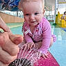 baby, child, infant, smiling, swimsuit, pink_clothing, water, splash_pad, swimming_pool, indoor_pool, water_play, fountain, slide, playground, adult, parent, support, sitting, portrait, happy