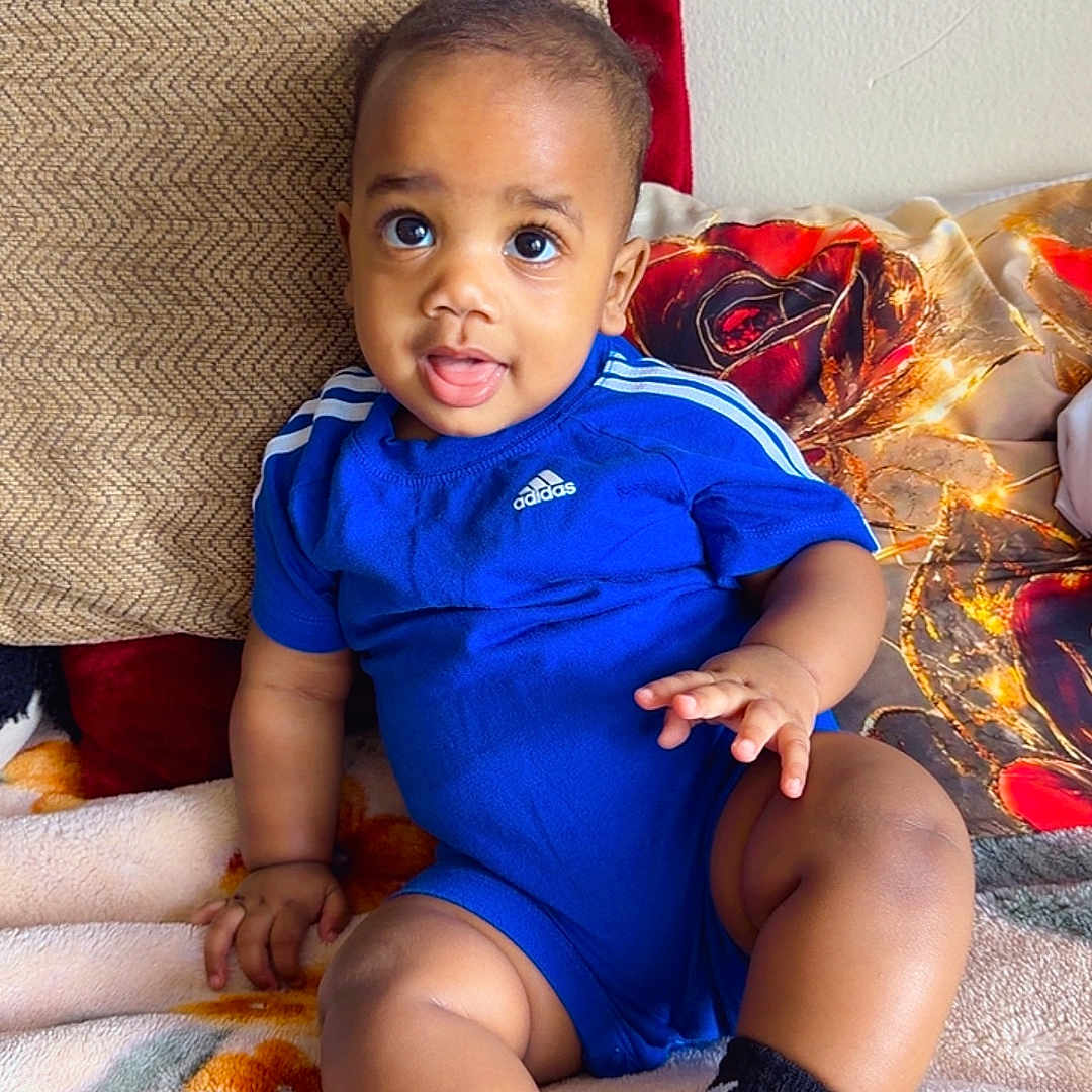 Rhaymond is registered to the contest to win money with this photo: baby, infant, child, blue_clothing, onesie, socks, sitting, blanket, pillow, floral_pattern, indoor, cute, face, portrait, smiling, looking_at_camera, skin, hands, legs, comfortable