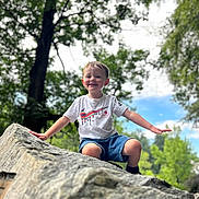 Ezra is registered to the contest to win money with this photo: child, boy, smiling, outdoor, rock, tree, nature, sky, greenery, tshirt, shorts, playful, happy, daylight, casual_clothing, person, portrait, fun, balance, summer
