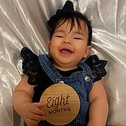 Aurora joined the competition — help win amazing prizes! baby, infant, smiling, denim_overalls, black_clothing, lace, headband, bow, wooden_sign, eight_months, hand, lying_down, white_satin_background, cute, happy, portrait, child, skin, face, person