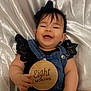 baby, infant, smiling, denim_overalls, black_clothing, lace, headband, bow, wooden_sign, eight_months, hand, lying_down, white_satin_background, cute, happy, portrait, child, skin, face, person