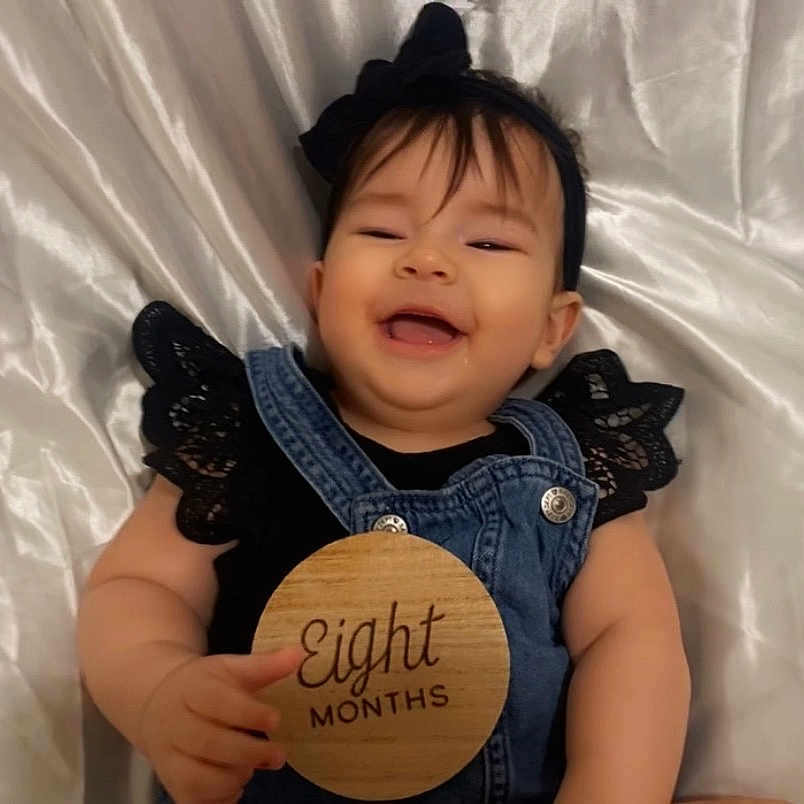 Aurora joined the competition — help win amazing prizes! baby, black_clothing, bow, child, cute, denim_overalls, eight_months, face, hand, happy, headband, infant, lace, lying_down, person, portrait, skin, smiling, white_satin_background, wooden_sign